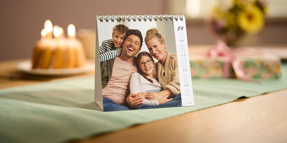 Ein CEWE Fotokalender im Tischformat zeigt im April ein Familien-Porträt und steht dekorativ auf einem festlich gedeckten Tisch.