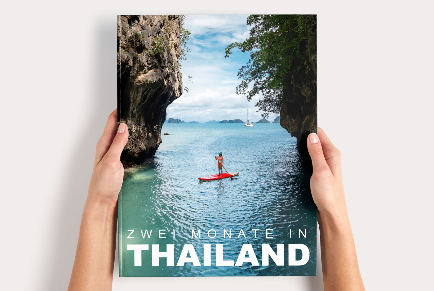 Hände halten ein CEWE FOTOBUCH XXL mit Stand-up-Paddler in türkisblauer Bucht zwischen Felsen, Titel "ZWEI MONATE IN THAILAND"