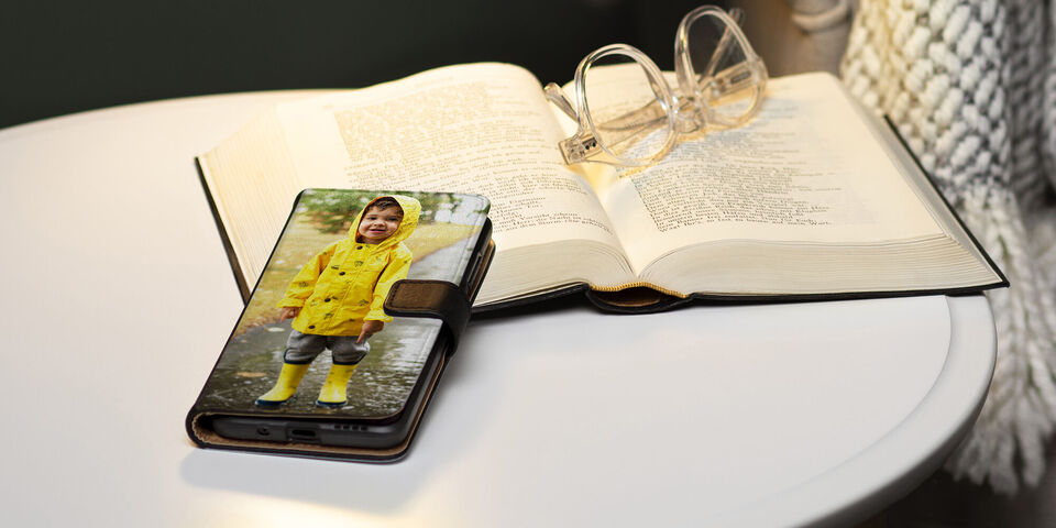 Ein Smartphone auf einem Tisch zeigt ein fröhliches Kind im Regenmantel und Gummistiefeln. Neben dem Telefon liegt ein aufgeschlagenes Buch und eine Brille.
