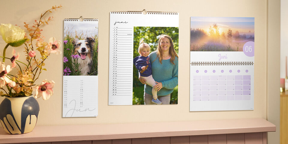 Drei CEWE Foto-Terminkalender hängen nebeneinander an einer Wand. Ein Hundefoto, ein fröhlicher Outdoor‑Moment und eine Morgenlandschaft.