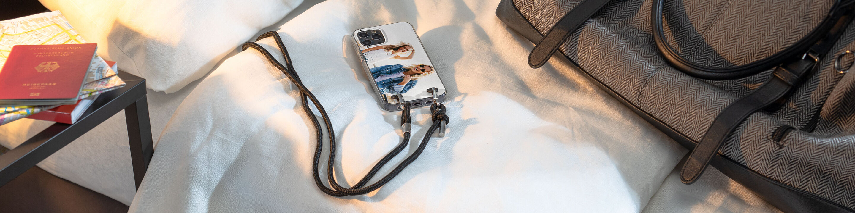 Ein Handy mit einem personalisierten Cover auf einem Bett neben einer Tasche und einem Reisepass.