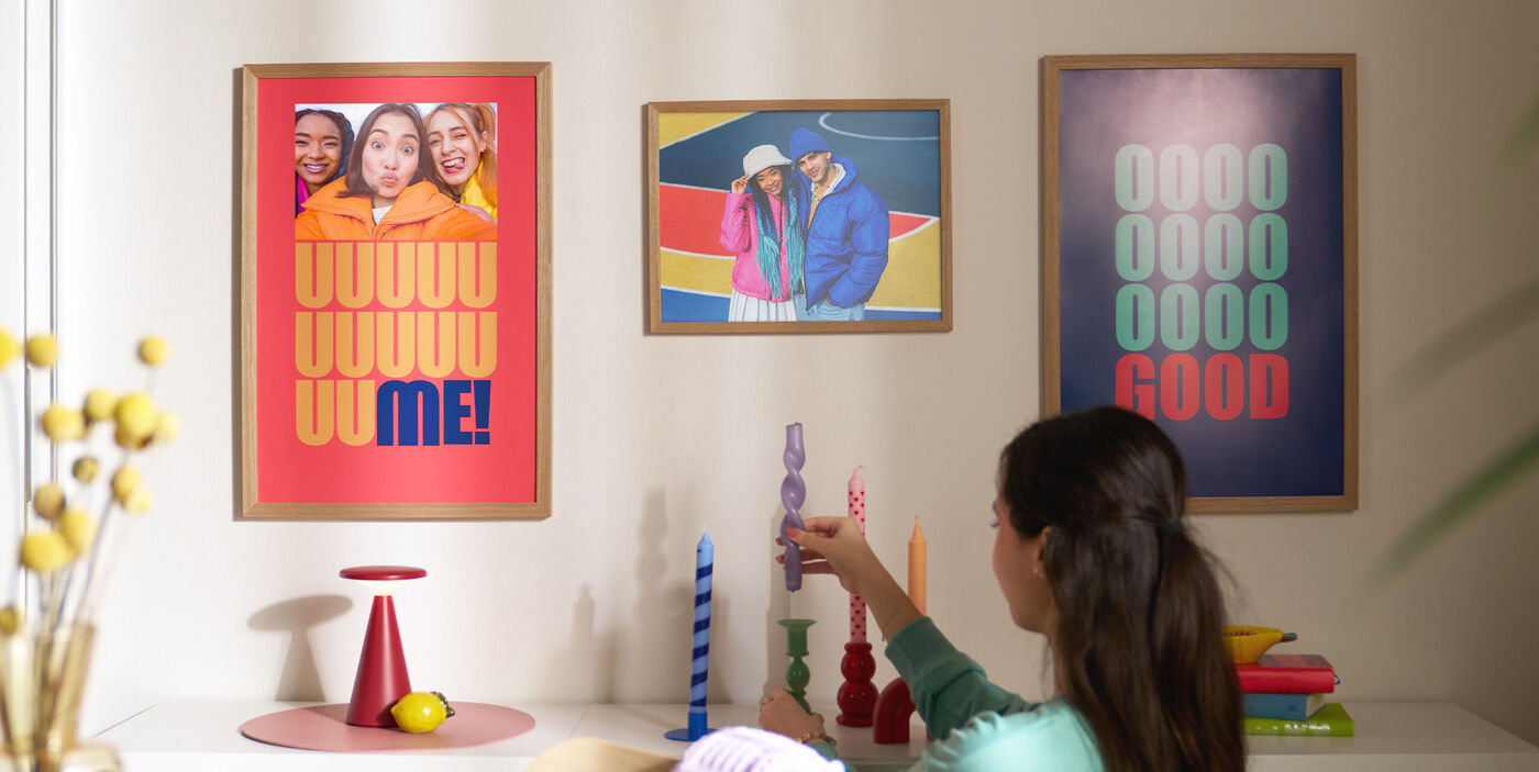 Eine Frau dekoriert ein Wohnzimmerregal. An der Wand hängen drei Poster, die mit Fotos und Schriftzügen gestaltet sind.