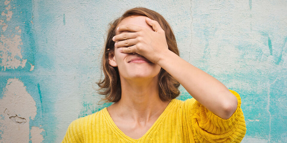 Junge Frau mit Hand vor dem Gesicht in gelbem Pullover vor einer blauen Wand.
