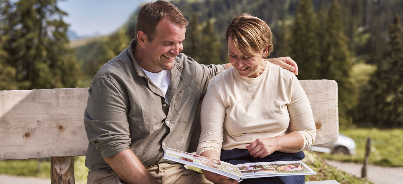Ein Paar betrachtet gemeinsam ein CEWE FOTOBUCH auf einer Bank in der Natur.