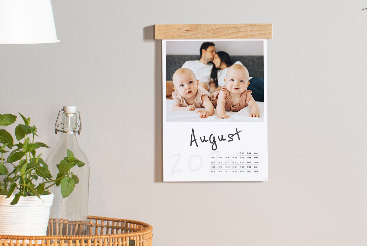 Ein Kalenderblatt für August zeigt glückliche Kinder auf einem Bett, die auf den Betrachter schauen. Im Hintergrund sind Eltern zu sehen, die sich umarmen