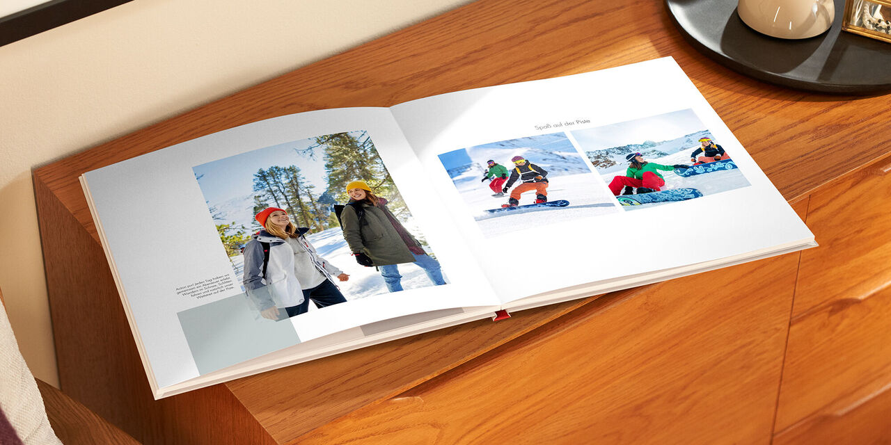 Auf einem Holzschrank liegt das aufgeklappte Fotobuch. Links sieht man ein Foto von zwei Frauen bei einer Wanderung. Daneben befindet sich ein kleiner Textblock. Auf der rechten Seite sind zwei Fotos der Frauen beim Snowboarden zu sehen. Darüber steht die Überschrift «Spass auf der Piste».