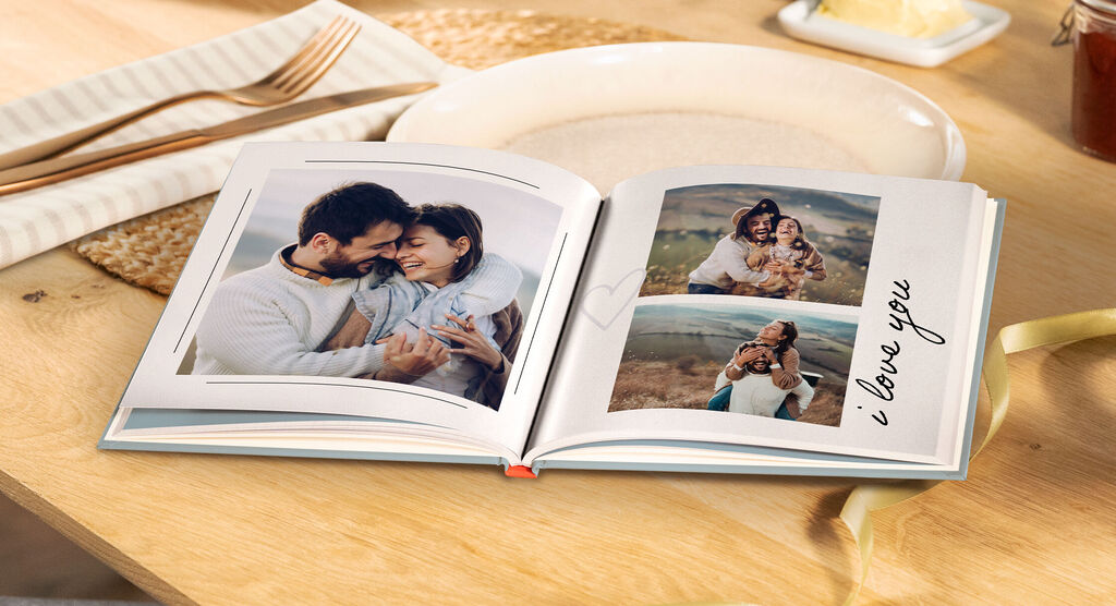 Ein offenes CEWE FOTOBUCH mit liebevollen Bildern von einem Paar und einer Familie. Der Text "I love you" ist neben einem der Fotos zu sehen. Im Hintergrund sind ein Tisch und eine gedeckte Tafel zu erkennen.