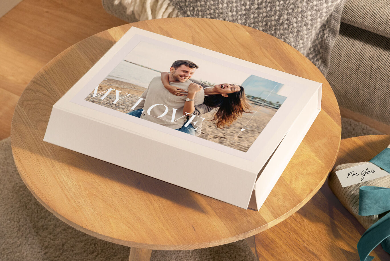 Ein Paar sitzt am Strand und strahlt vor Freude. Der Cover des CEWE FOTOBUCHS zeigt das Wort 'MY LOVE'.