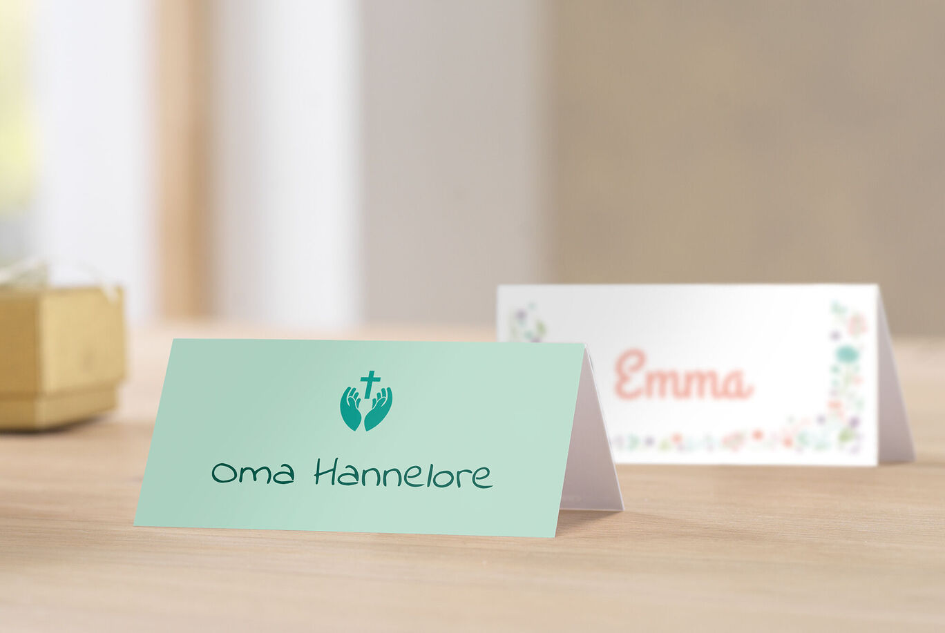 Zwei liebevoll gestaltete Tischkarten für besondere Anlässe. Eine Karte in sanftem Grün mit einem Kreuz und dem Namen "oma Hannelore" darauf, die andere in weiß mit floralen Elementen und dem Namen "Emma".