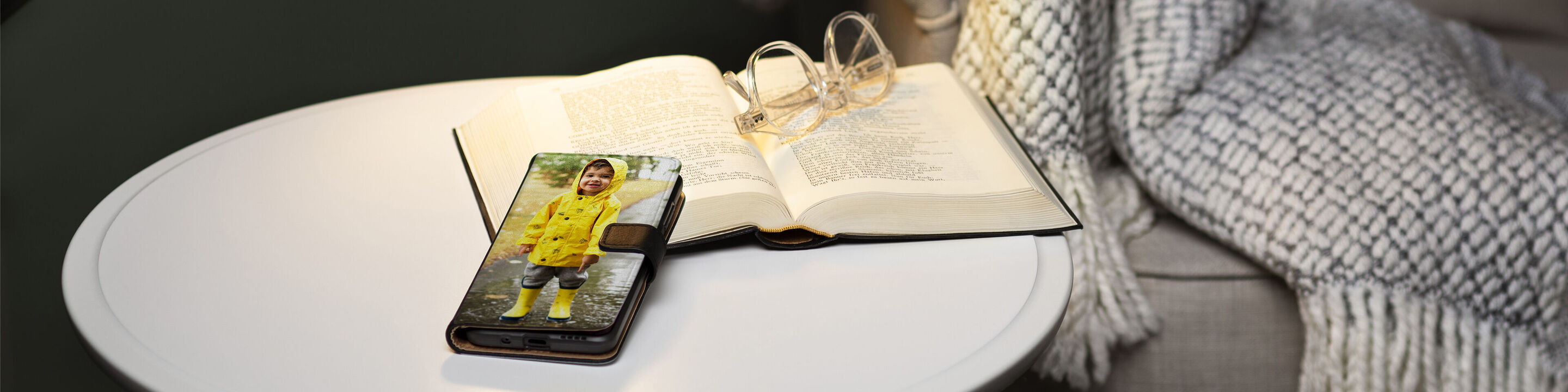 Ein Smartphone mit einem Bild eines Kindes in einem gelben Regenmantel auf einem Tisch, neben einem offenen Buch und einer Brille.