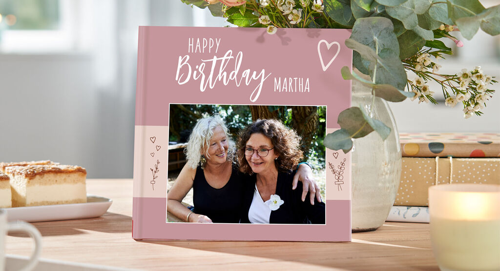 Zwei Freundinnen feiern einen Geburtstag mit einem CEWE FOTOBUCH neben einer schönen Blumenarrangierung