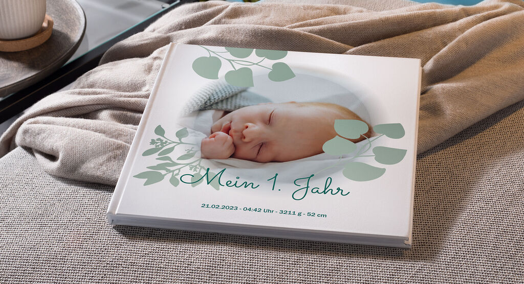 Ein CEWE FOTOBUCH mit dem Titel 'Mein 1. Jahr' auf einem Sofa, umgeben von sanften Farben und Naturmotiven