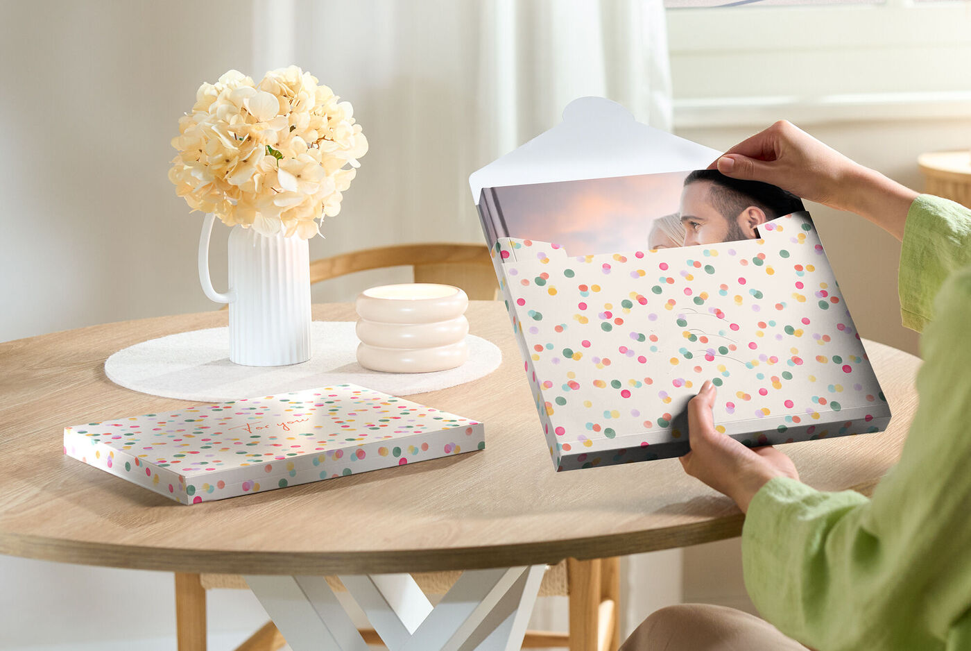 Eine Person öffnet ein CEWE FOTOBUCH mit einem fröhlich gestalteten Cover auf einem Tisch. Ein Blumenstrauß und weitere Deko ergänzen die Szene.