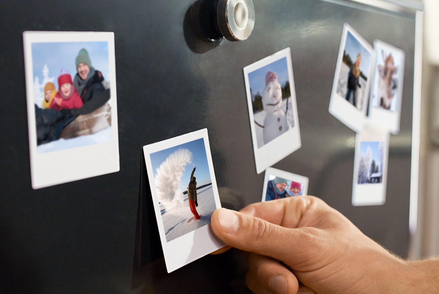 Eine Hand befestigt einen Retro‑Fotomagneten an einer magnetischen Oberfläche, umgeben von weiteren winterlichen Fotomagneten.