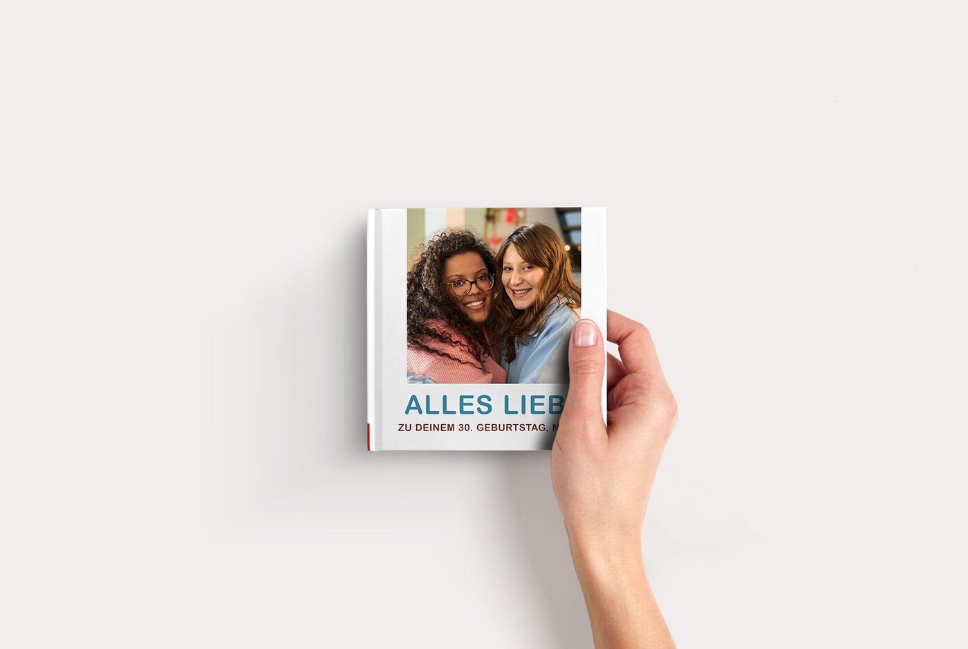 Hand hält ein CEWE Fotobuch Compact Quadratisch mit zwei Personen, die sich umarmen, und Schriftzug "ALLES LIEBE"