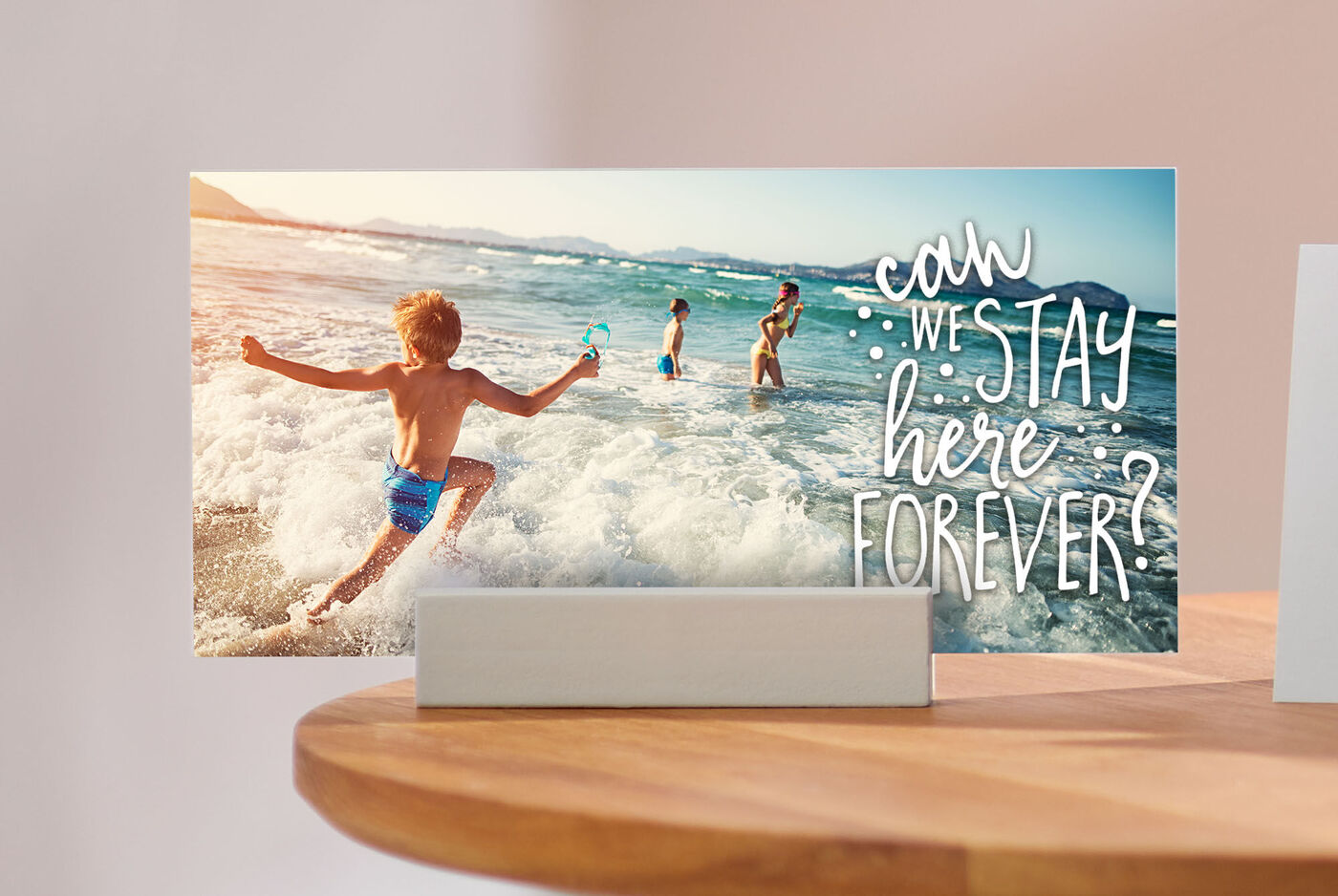 Ein Junge springt fröhlich in die Wellen der Küste, während andere Kinder im Wasser spielen. Auf dem Bild steht der Text: 'Can we stay here forever?'