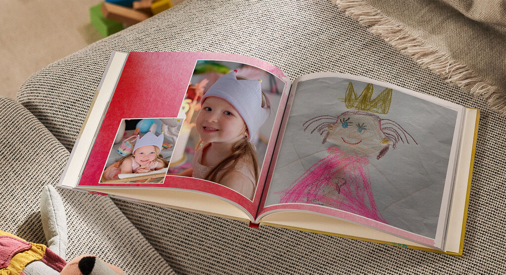 Ein offenes CEWE FOTOBUCH mit bunten Seiten. Eine handgezeichnete Figur mit einer Krone ist darauf zu sehen, sowie ein Foto eines lächelnden Kindes. Das CEWE FOTOBUCH strahlt Freude und Kreativität aus