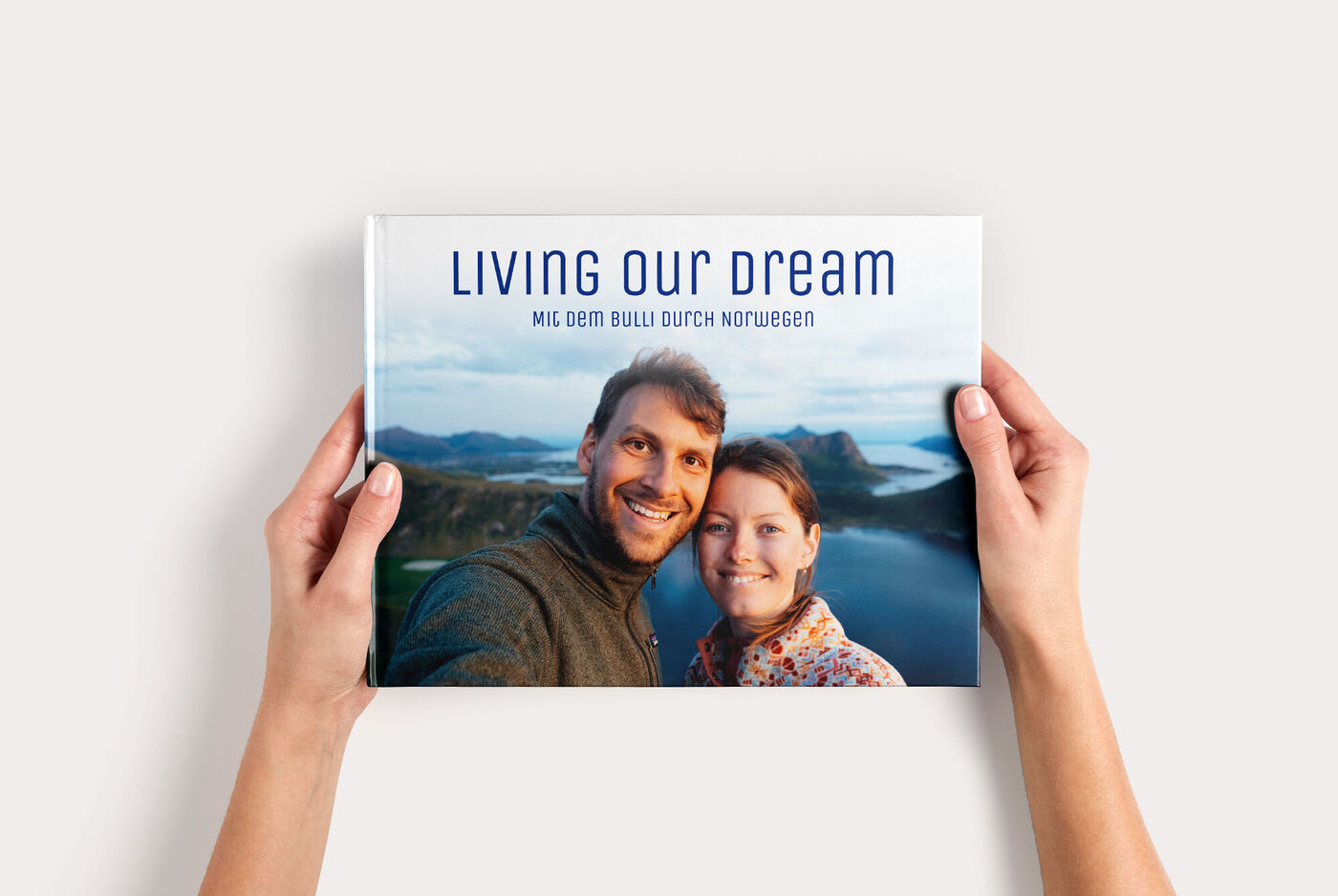 Zwei Hände halten ein CEWE FOTOBUCH Groß Panorama mit Paarfoto vor Bergsee/Fjord und Titel "LIVING OUR DREAM"
