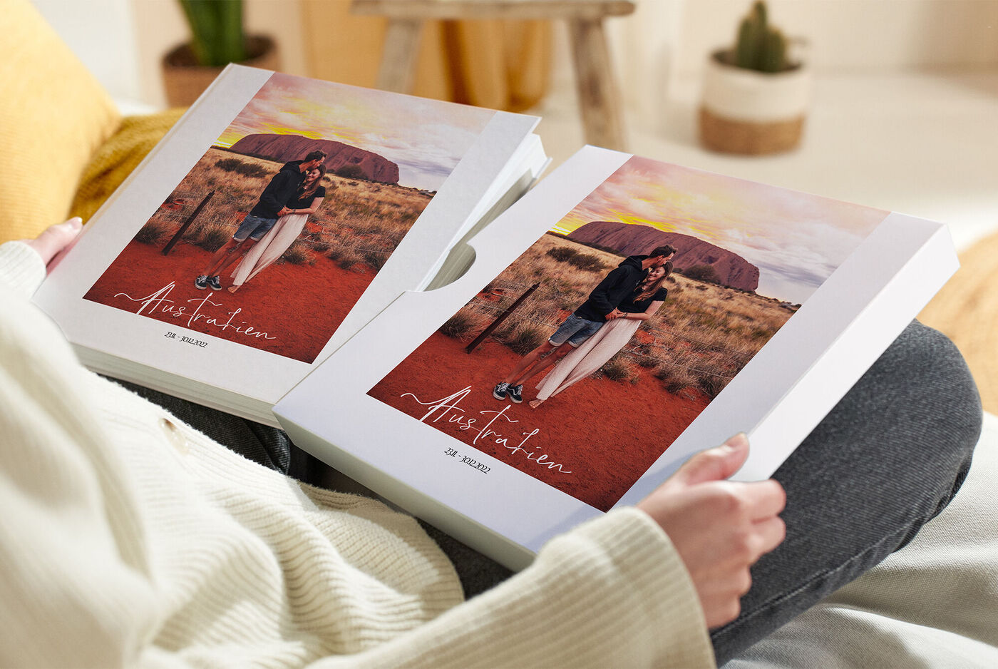 Eine Person hält zwei CEWE Fotobücher mit einem farbenfrohen Cover auf dem Schoß. Der Hintergrund zeigt eine Landschaft mit einem roten Erdboden und einem warmen Sonnenuntergang.