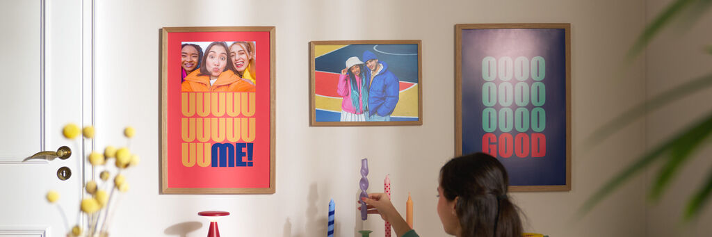 Eine Frau dekoriert ein Wohnzimmerregal. An der Wand hängen drei Poster, die mit Fotos und Schriftzügen gestaltet sind.